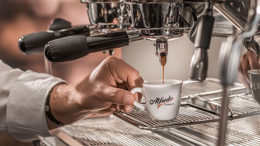 Barista zet een verse espresso in een stijlvol Alfredo kopje bij een koffiemachine.
