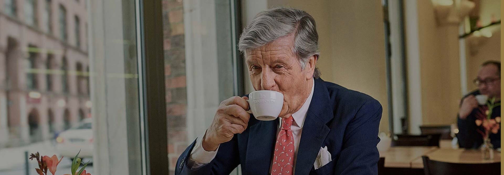 Wyrafinowany mężczyzna delektujący się kawą w kawiarni, otoczony eleganckim wystrojem i widokiem na ulicę miasta.