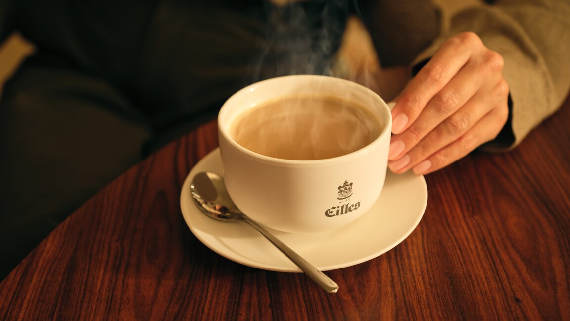 Eine dampfende Tasse Eilles-Kaffee steht auf einem Holztisch, eine Hand hält sie sanft.