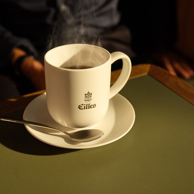 Eine dampfende Tasse Eilles-Kaffee auf einer Untertasse mit einem Löffel, die auf einer grünen Tischplatte in einer gemütlichen Umgebung steht.