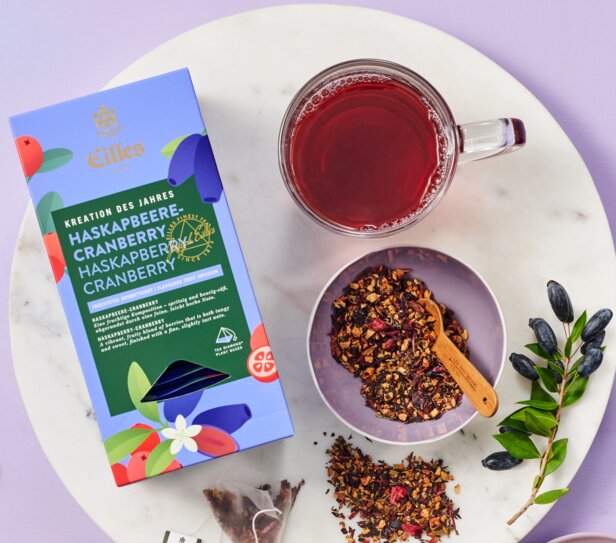 Lebendige Eilles-Tee-Verpackung mit Cranberry-Aufguss, Trockenfrüchten und einer dampfenden Tasse auf einer stilvollen Marmoroberfläche.