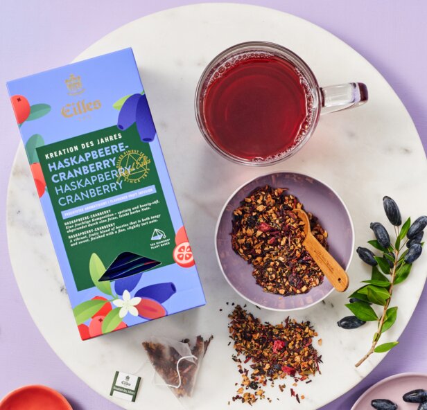 Vibrant Eilles tea packaging with cranberry infusion, dried fruits, and a steaming cup on a stylish marble surface.