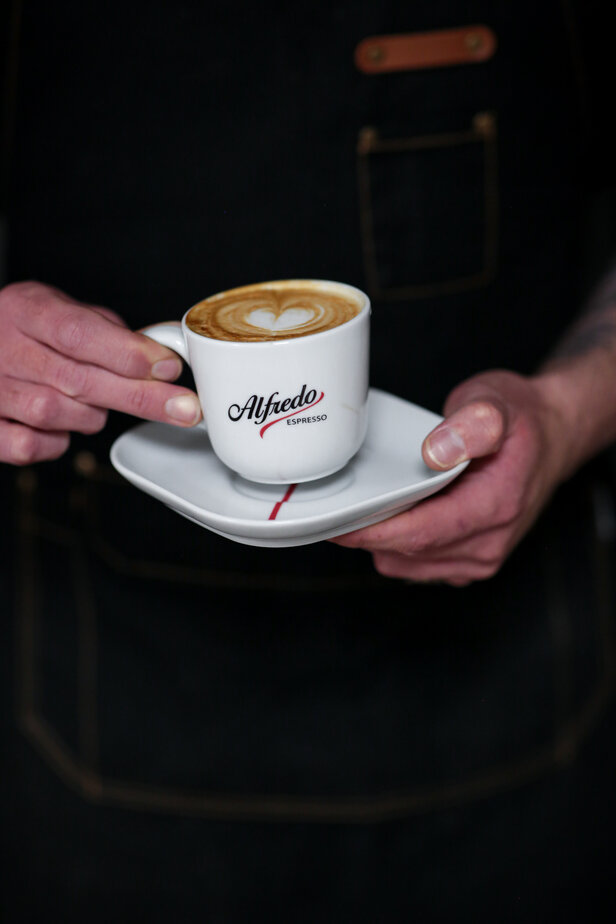 Barista presenting a latte art heart in a coffee cup from Alfredo Espresso on a white saucer.