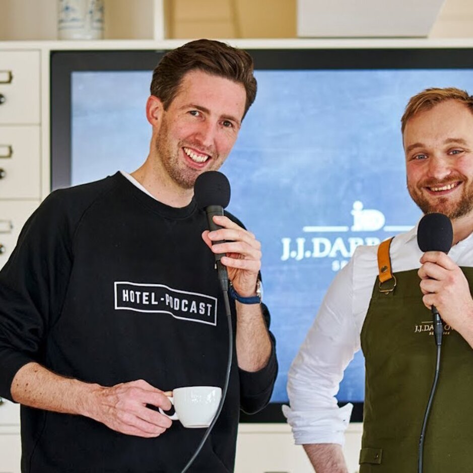 Podcast-Moderatoren diskutieren im stilvollen Ambiente des J.J. DARBOV Hotels mit Mikrofonen und Kaffeetassen über Trends im Gastgewerbe.