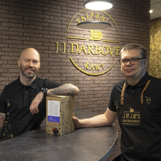 Dwóch baristów z dumą prezentuje kawę parzoną na zimno J.J. Jay w nowoczesnej kawiarni z ceglaną ścianą w tle.