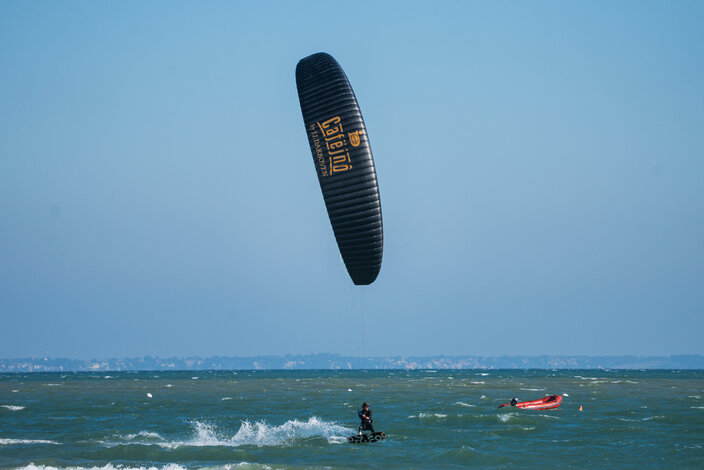 Kitesurfer szybuje nad falami z dużym czarnym latawcem, na tle czystego błękitnego nieba i odległej linii brzegowej.