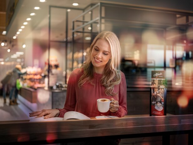 Kobieta pijąca kawę i czytająca książkę w modnej kawiarni, z menu zawierającym opcje espresso w tle.